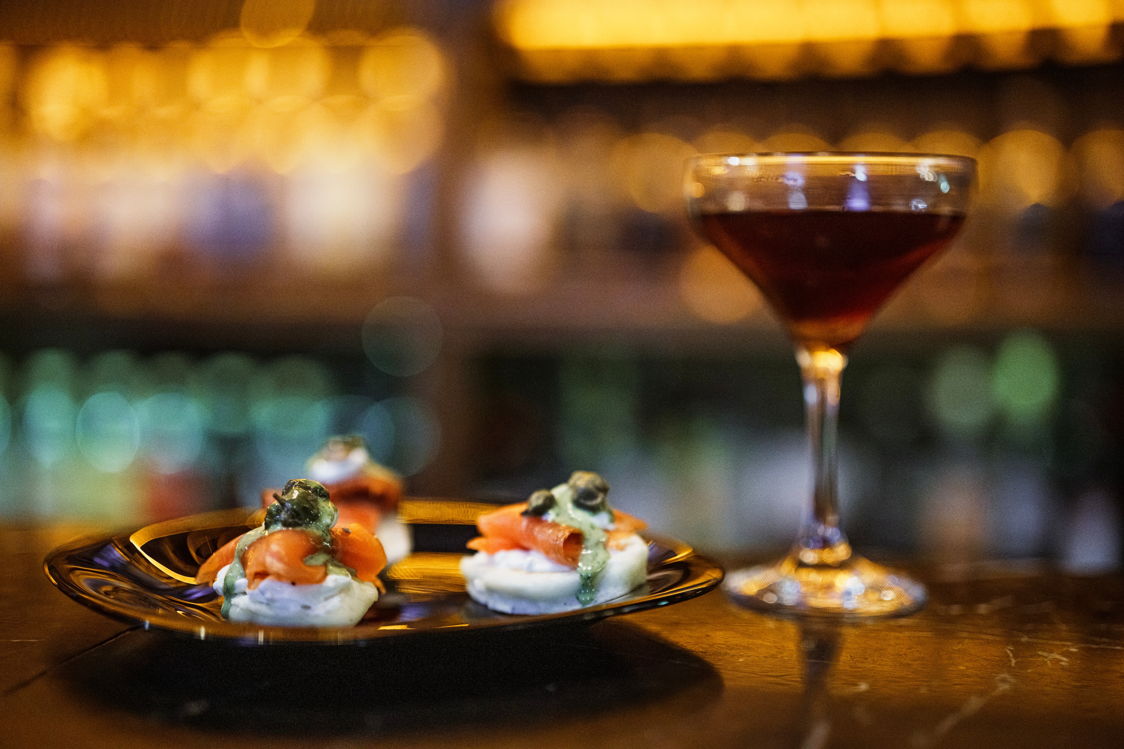 Elegant appetizers and a cocktail are displayed on a bar table with warm ambient lighting in the background.