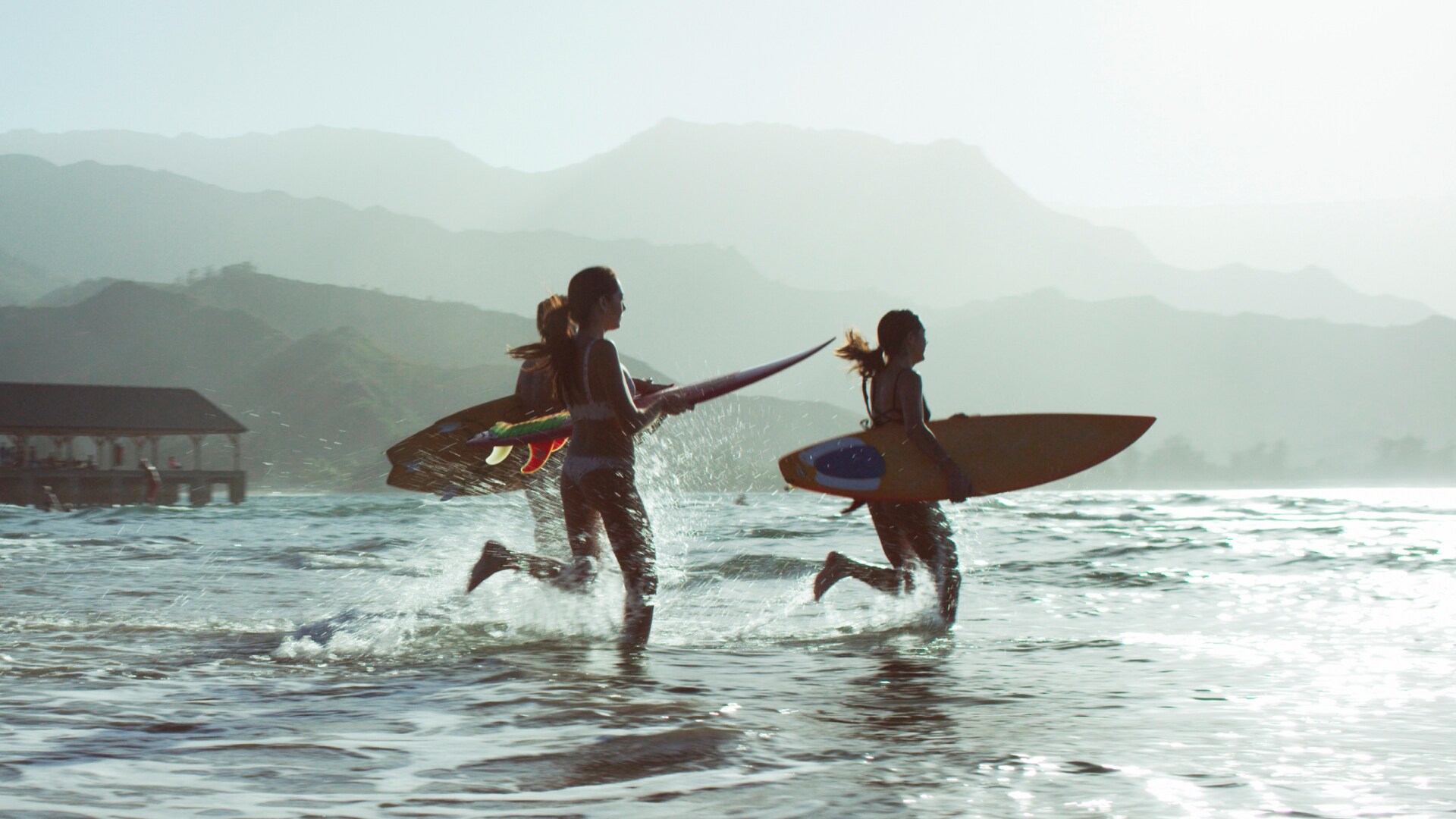 Surfers run into the ocean near a pier, Honolulu, Hawaii with mountains beyond
