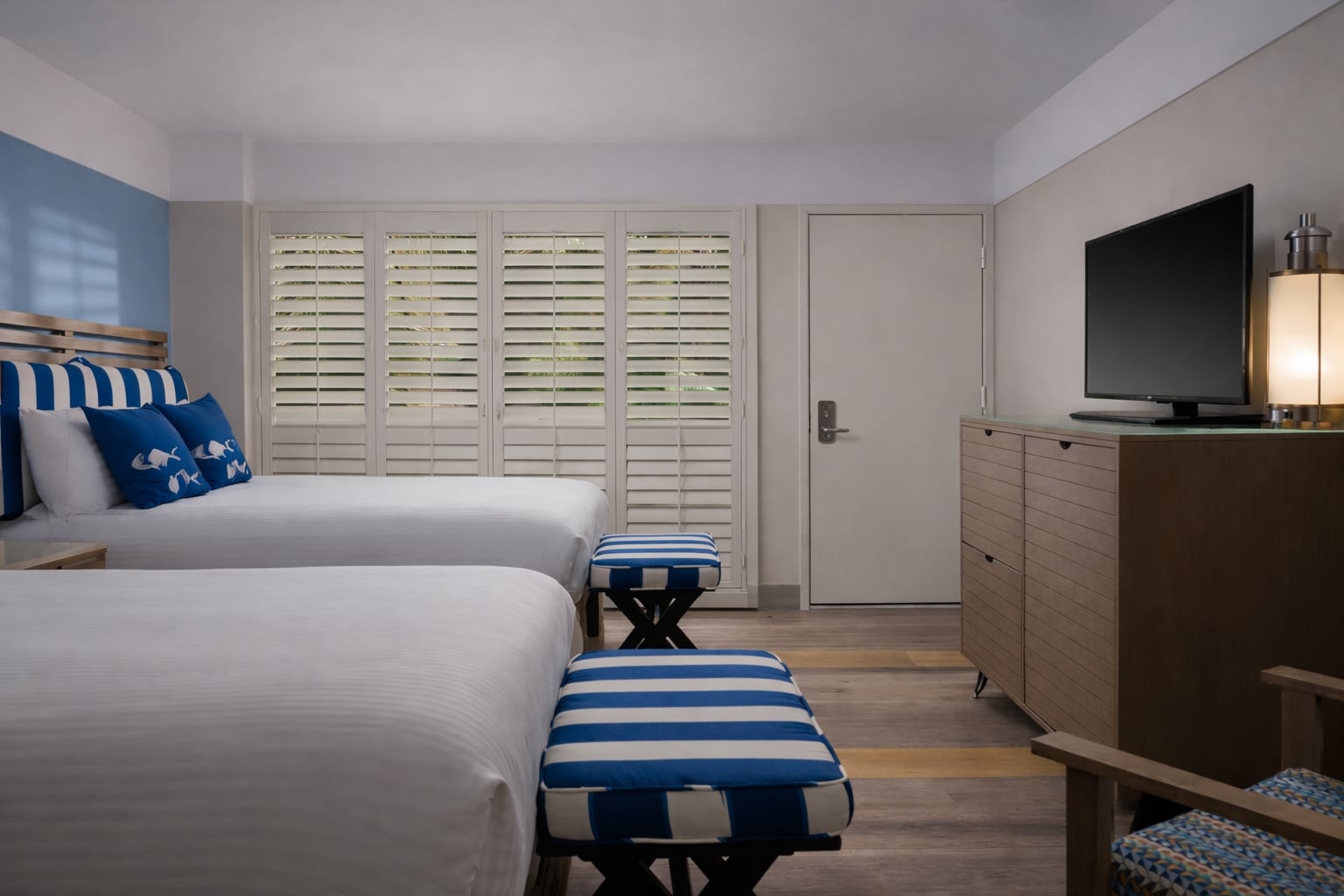 A hotel room with two beds, a TV on a dresser, and shutters on the windows.