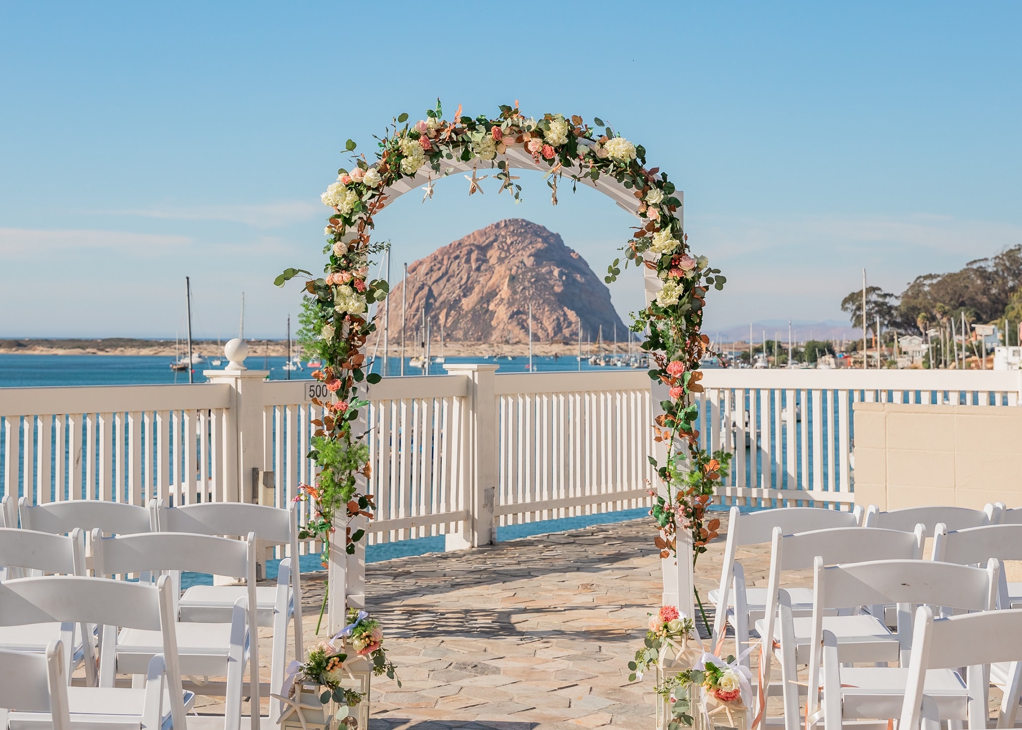 A floral arch with white chairs in front of a water view with a mountain in the background.