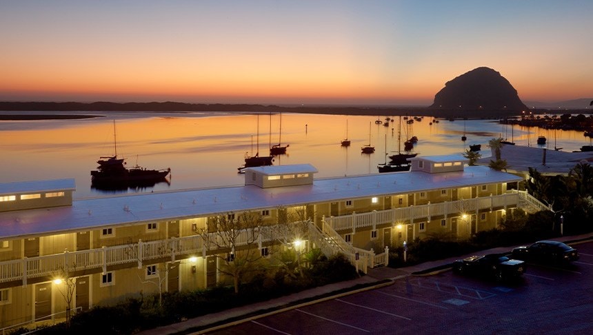 A two-story building with a white roof and yellow walls, overlooking a body of water with boats, at sunset.