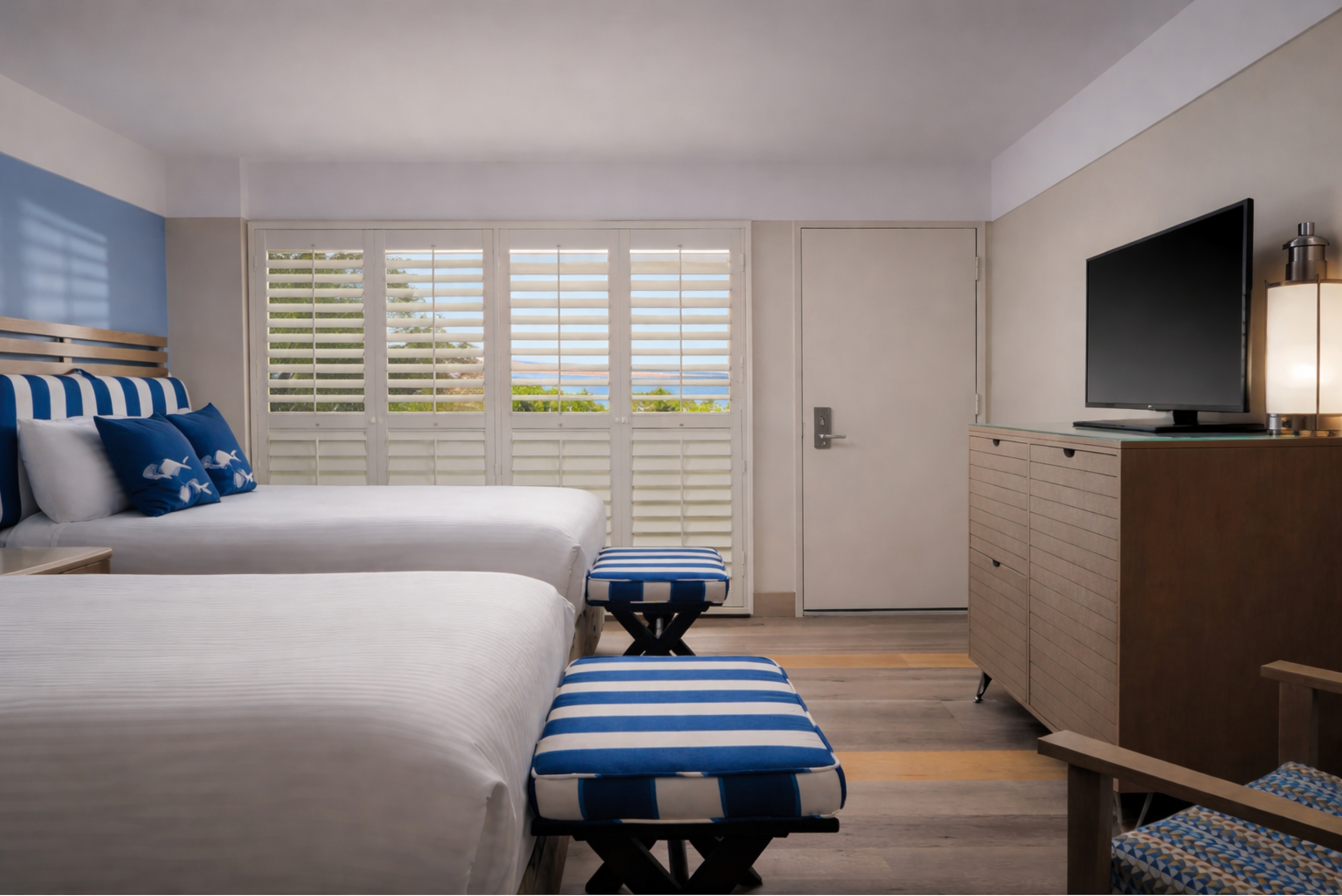 A hotel room with two twin beds, a window with blinds, a TV on a dresser, and two blue and white benches.