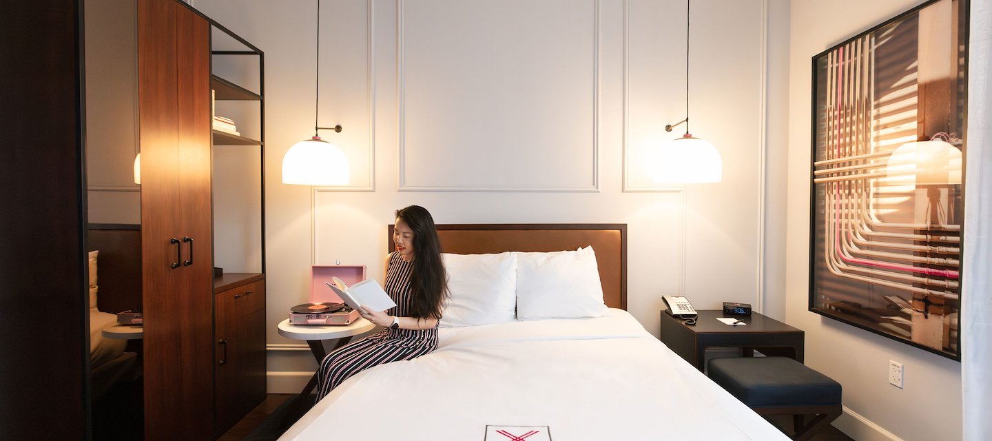 A young woman sitting on a king bed embroidered with the Axiom Hotel icon, reading a book and listening to a record