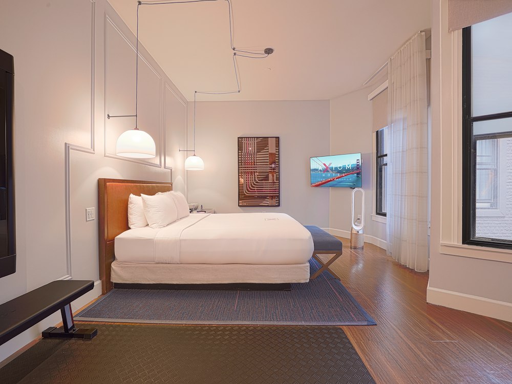 A wide view of a hotel room with a king-size bed, flat-screen TV, pendant lighting, wood floor, and large window.