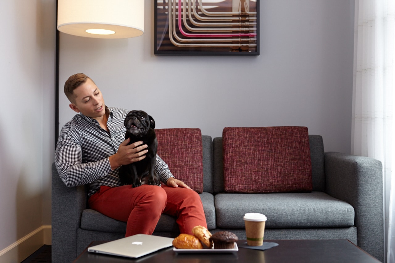 A man sitting on a couch with a dog, laptop, pastries, and coffee