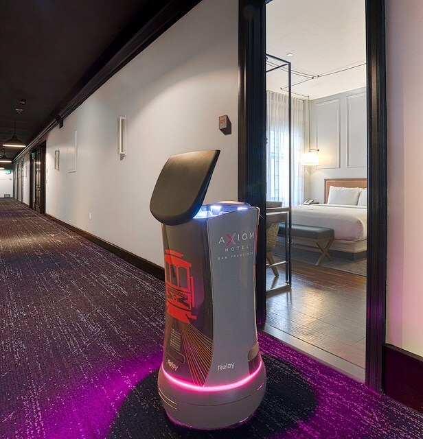 A robot stands in a hotel hallway with a doorway to a room.