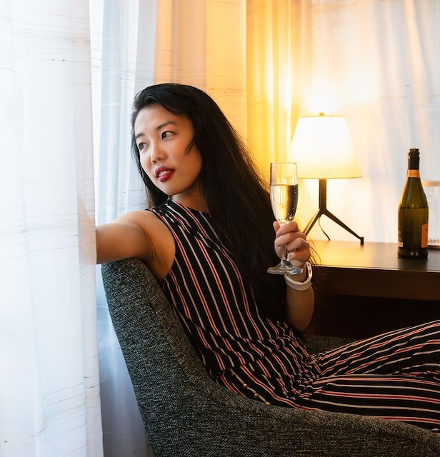 A woman sitting in a chair with a glass of champagne in a room with a table and lamp.