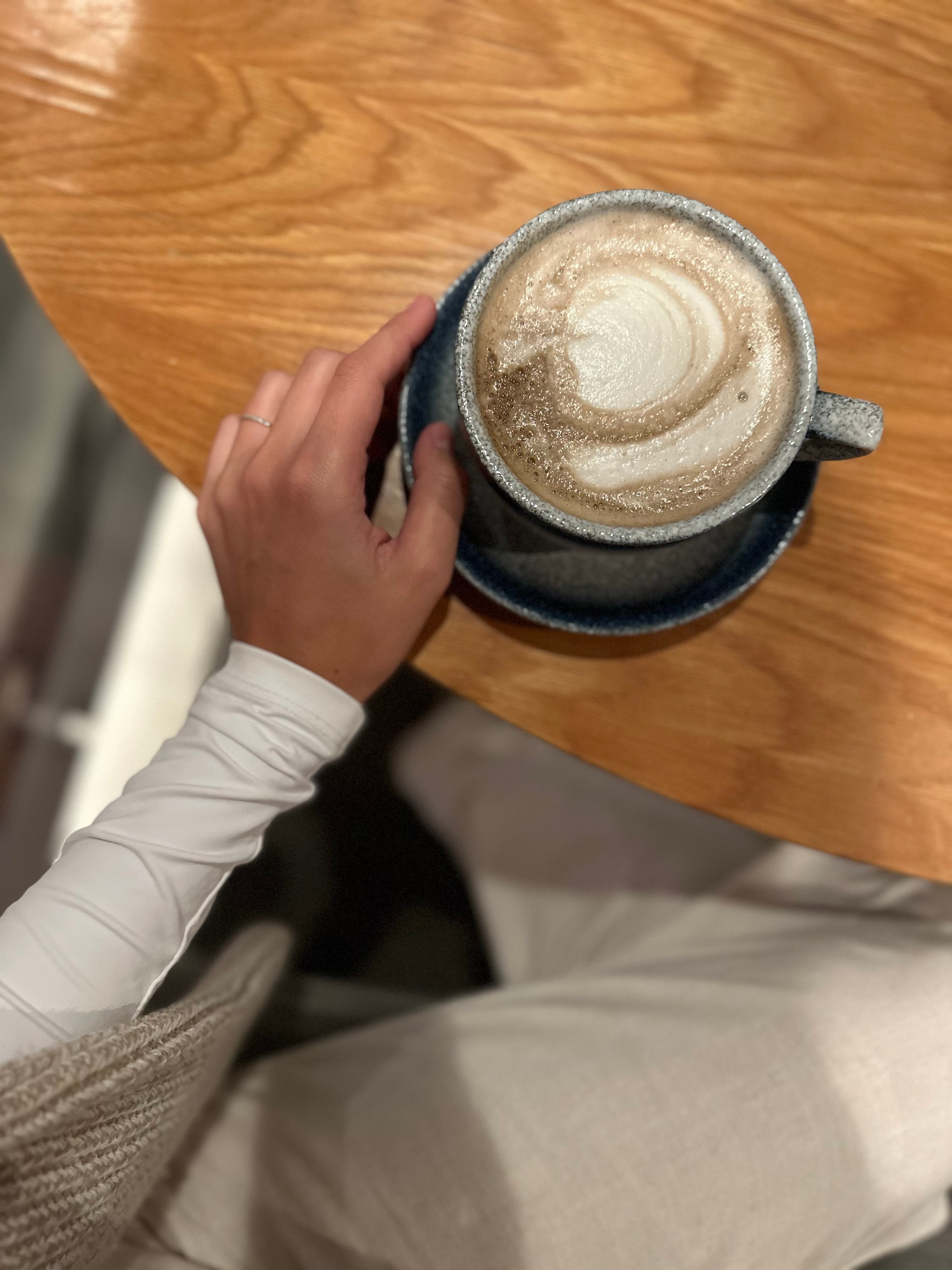 A hand holding a coffee cup on a saucer over a wooden table.