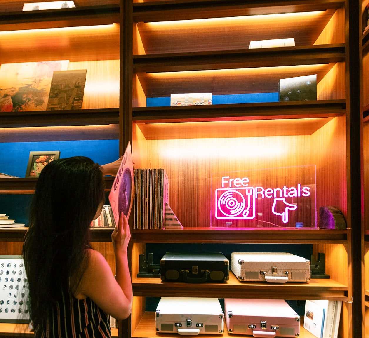 a woman browsing through records in front of a shelf with a neon sign that reads 'Free Vinyl Rentals'