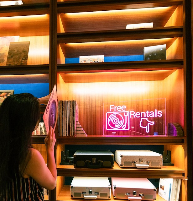 a woman browsing through records in front of a shelf with a neon sign that reads 'Free Vinyl Rentals'
