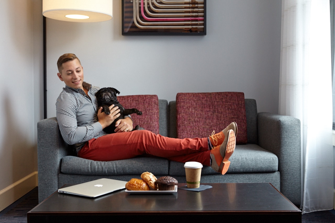 A man sitting on a sofa in a lobby lounge with a dog, a coffee table with snacks, and a laptop.