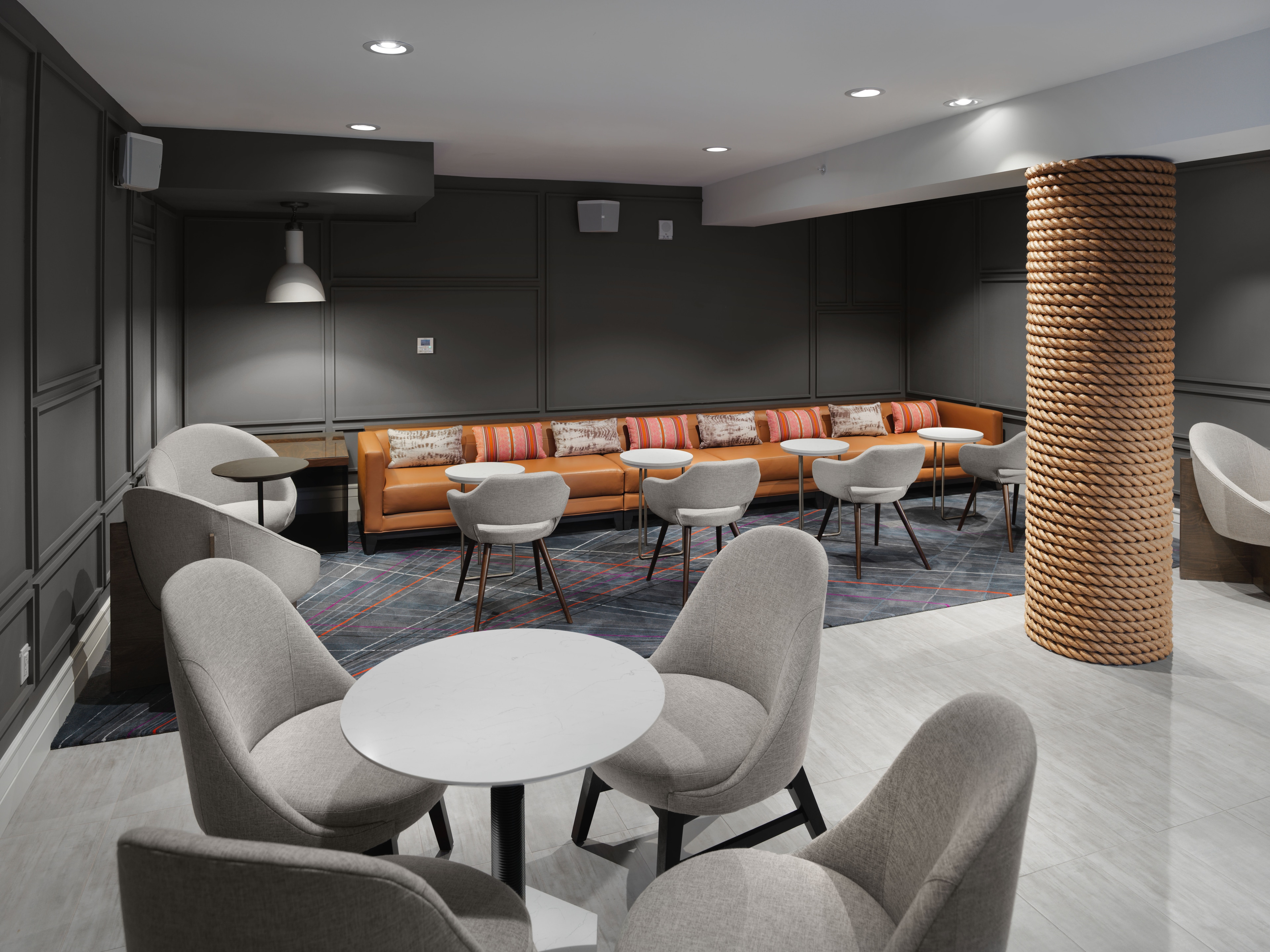 A modern lobby lounge with a long orange sofa, gray armchairs, and small tables.