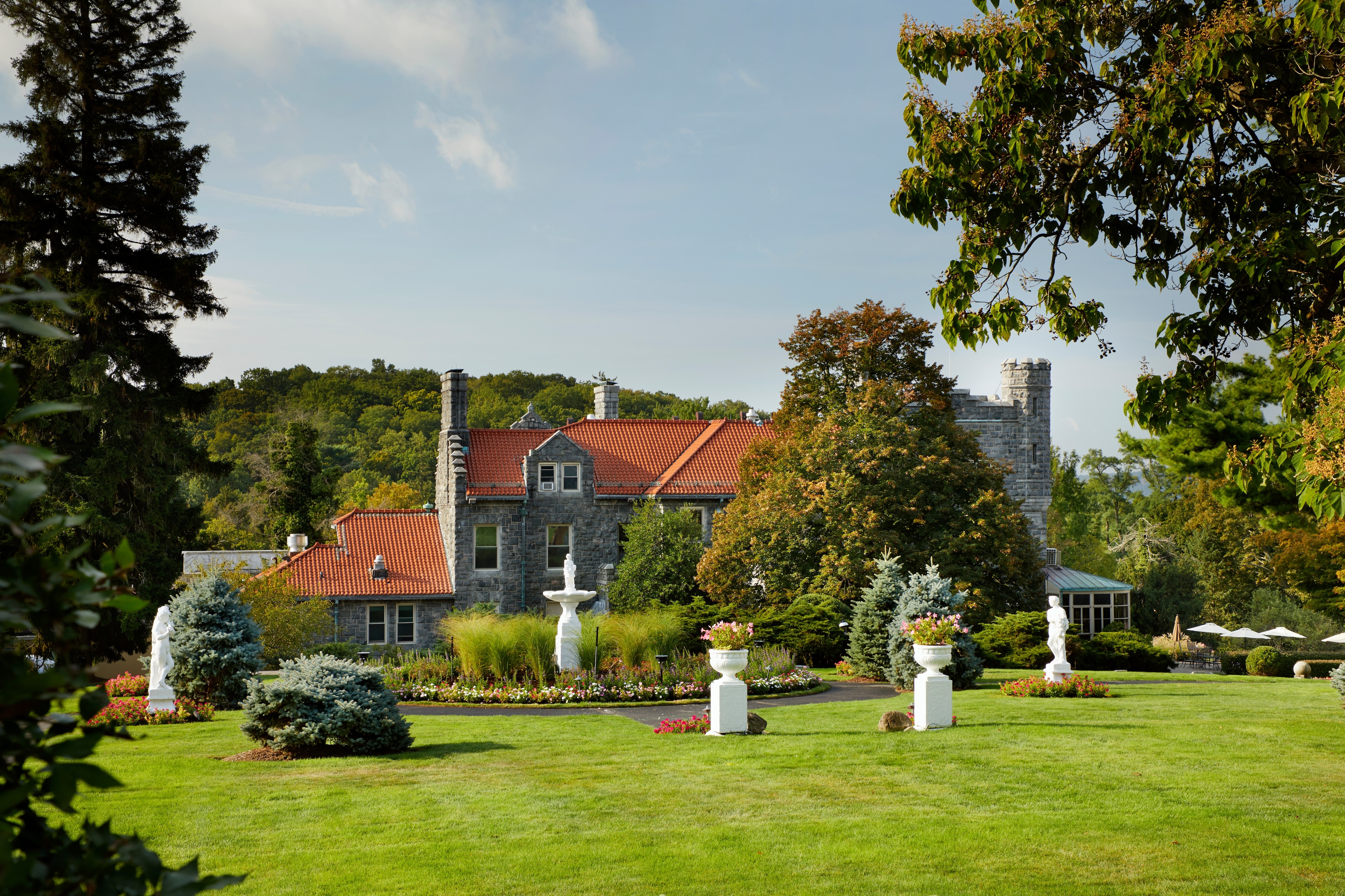 Historic stone mansion with red roof, surrounded by lush green gardens and tall trees