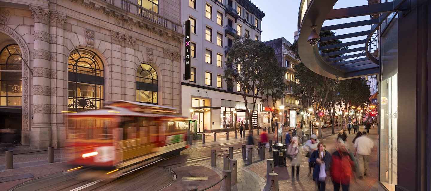 A wide exterior view of a hotel building with a stone facade, arched windows, and a vertical sign reading 'AXIOM' on a city street with a tram and pedestrians.