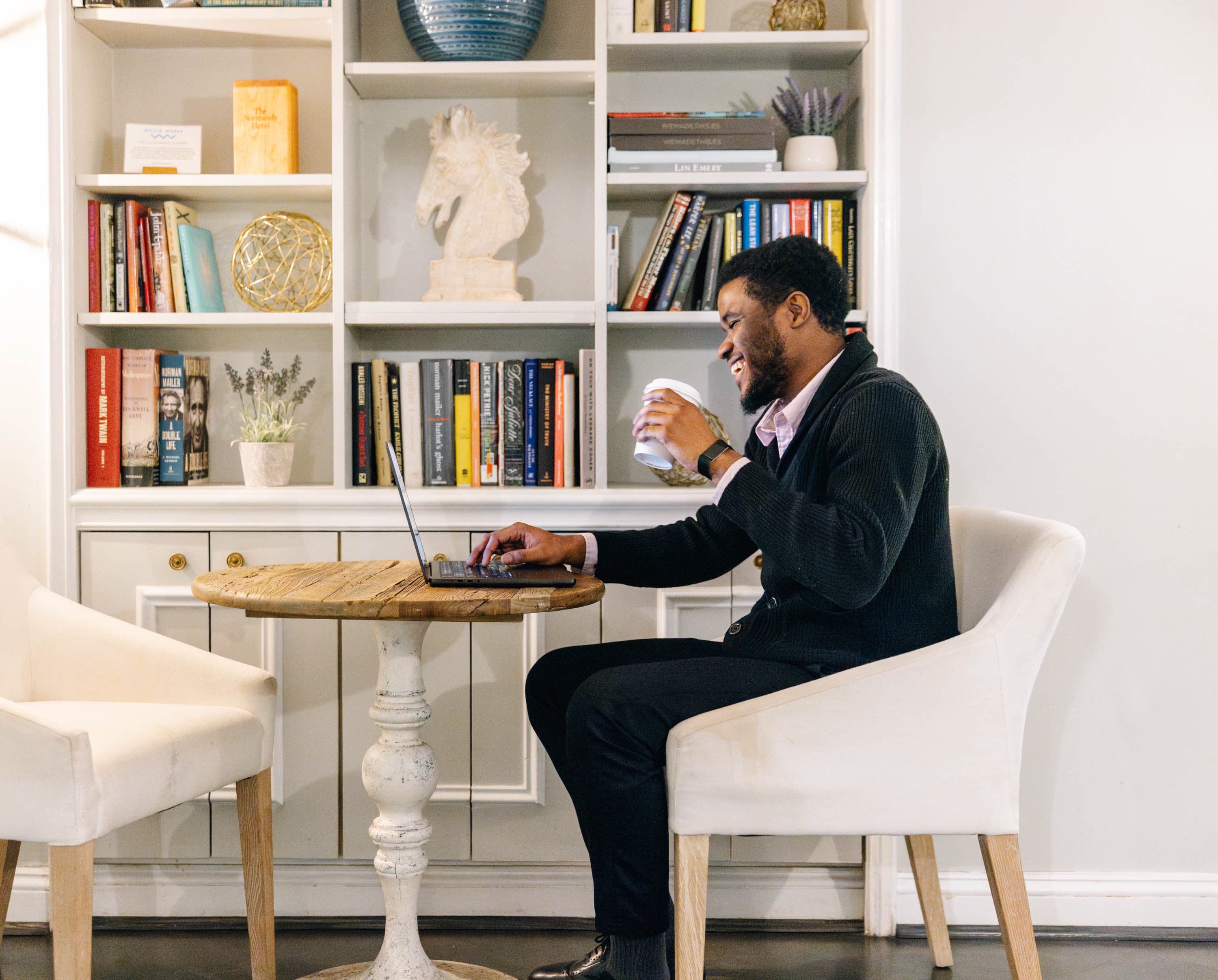A man sits at a small table in a lobby lounge, using a laptop and holding a cup.