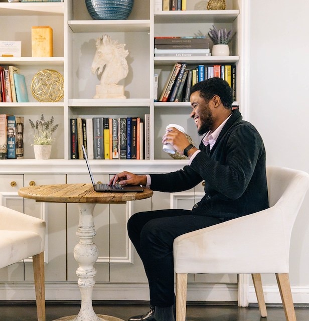 A man sits at a small table in a lobby lounge, using a laptop and holding a cup.
