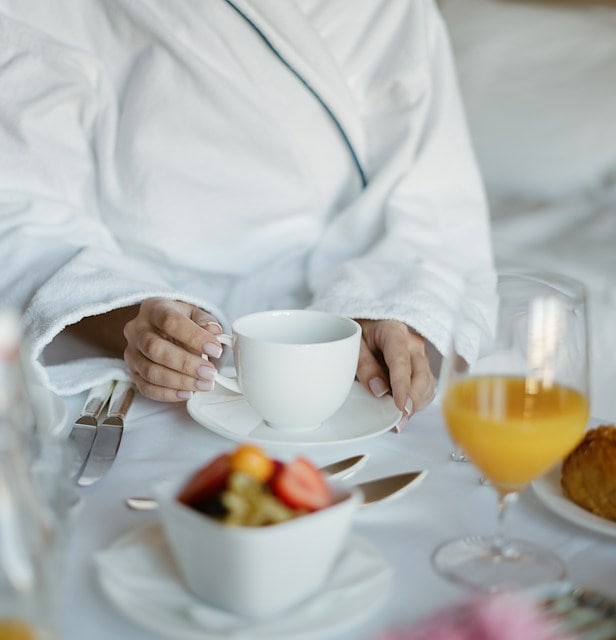 A person in a white robe holding a cup of coffee with a bowl of fruit and a glass of orange juice on a table.