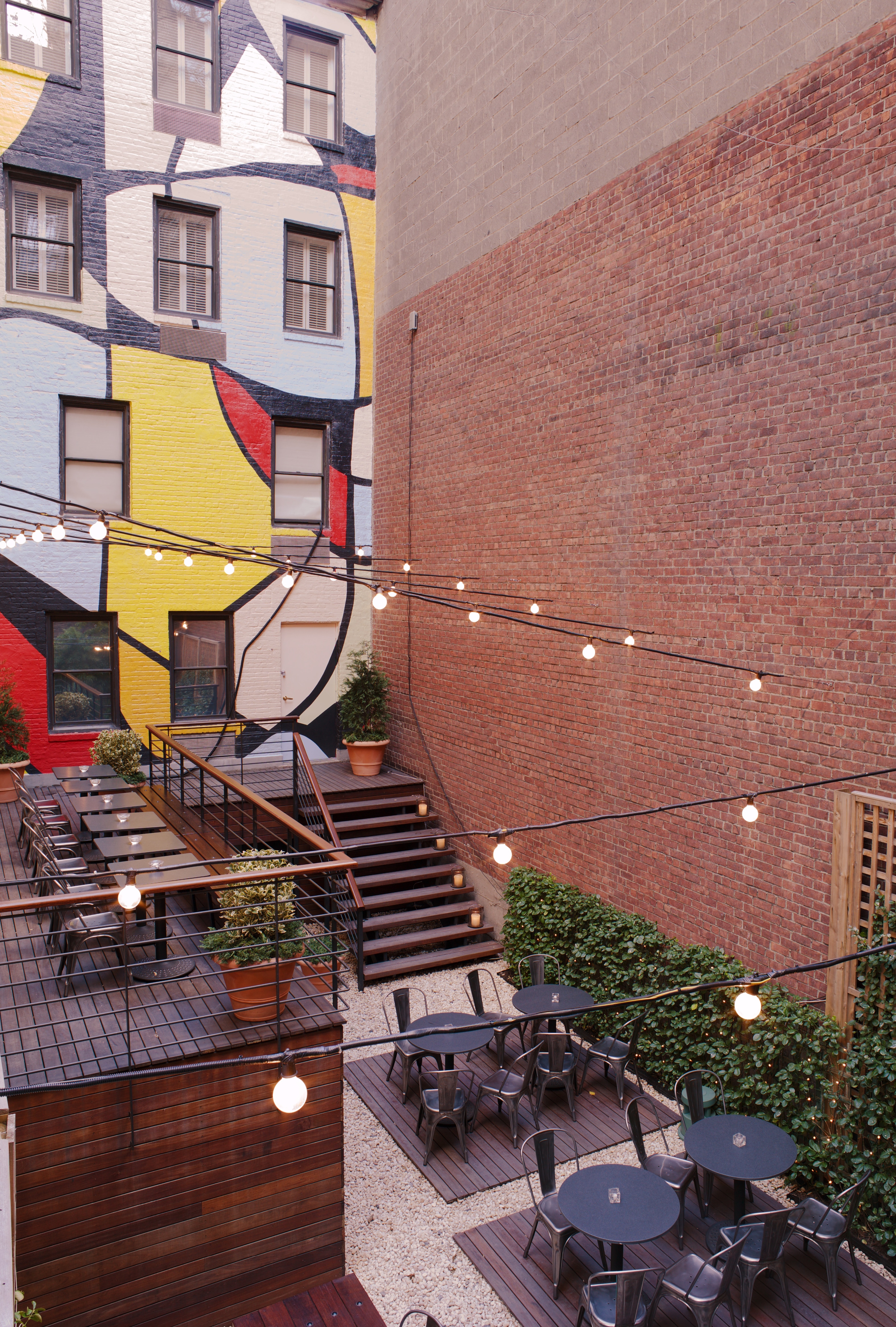 An outdoor seating area with tables and chairs, a mural on a building, and string lights.