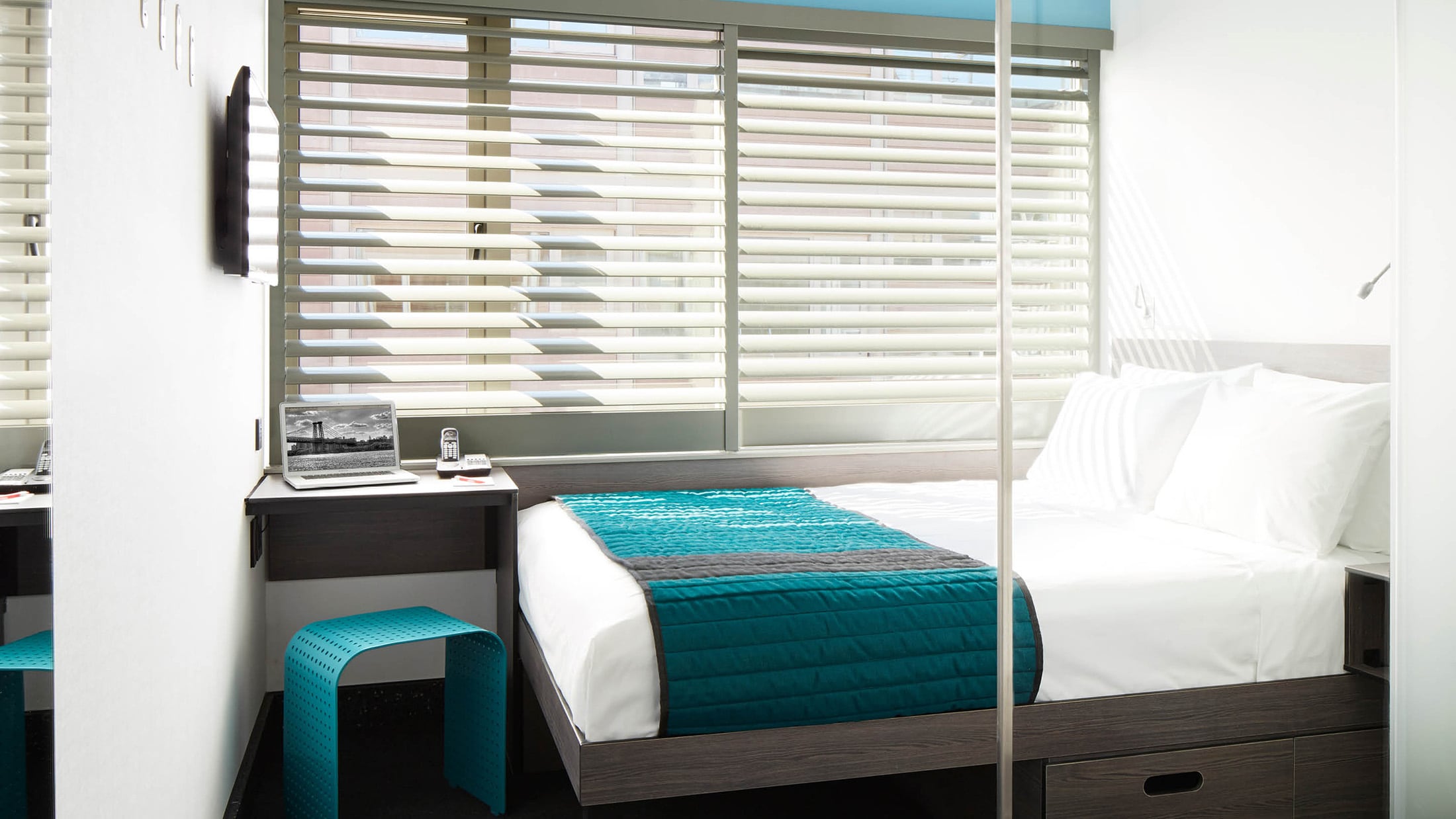 A hotel room with a bed, desk, and window covered in blinds.