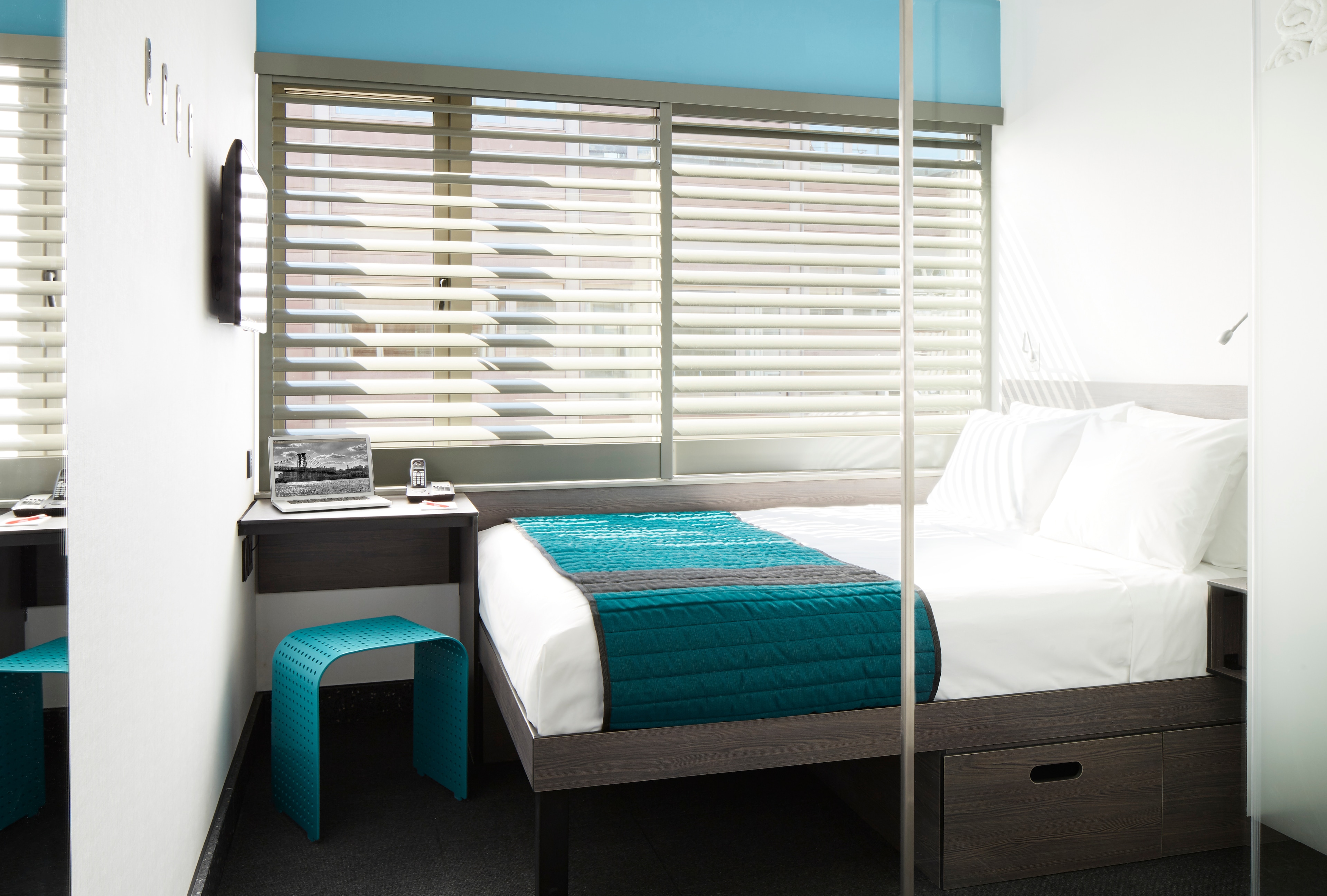A hotel room with a bed, desk, and window with blinds.