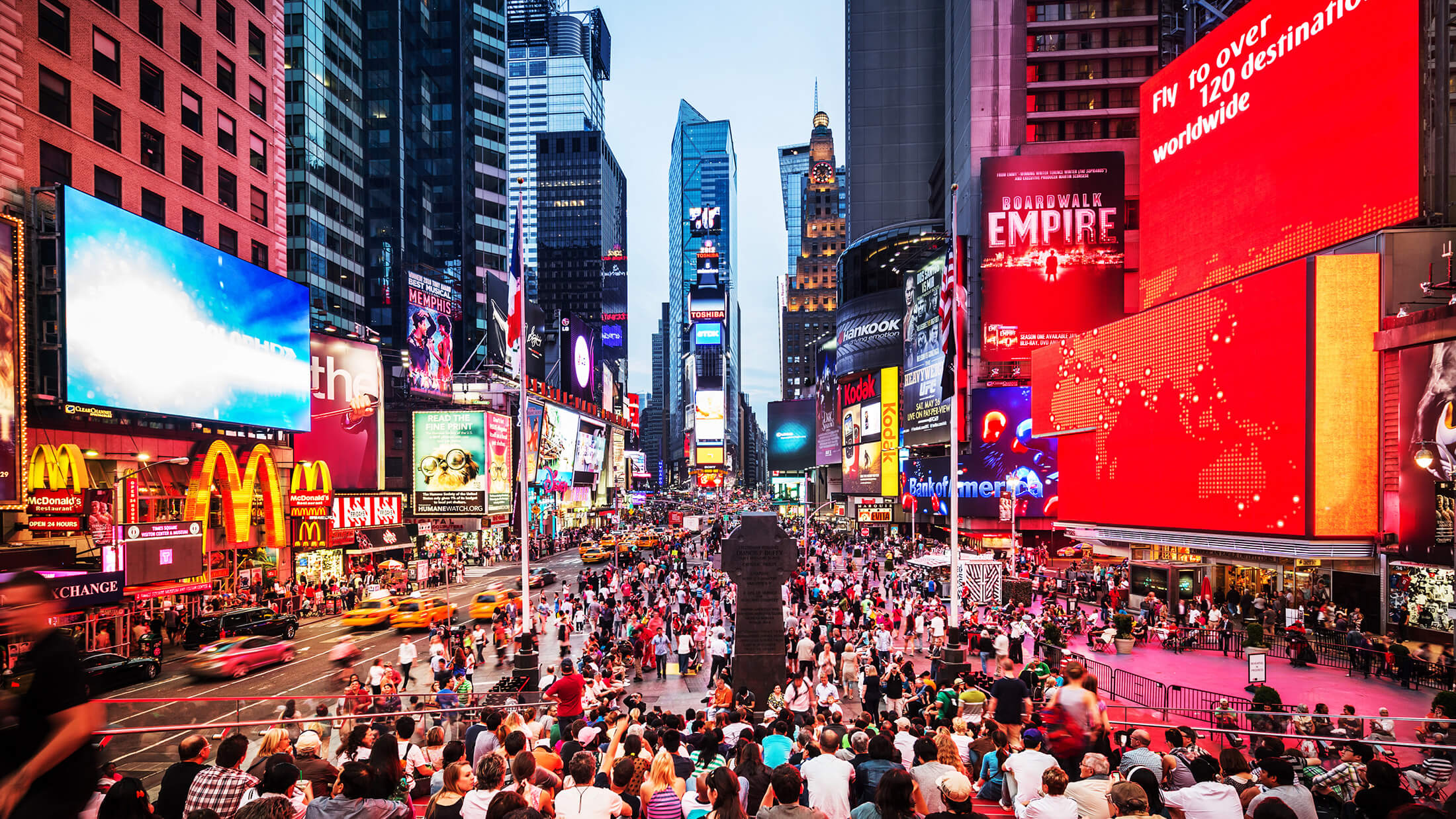 A bustling city street with tall buildings, bright billboards, and a large crowd of people.