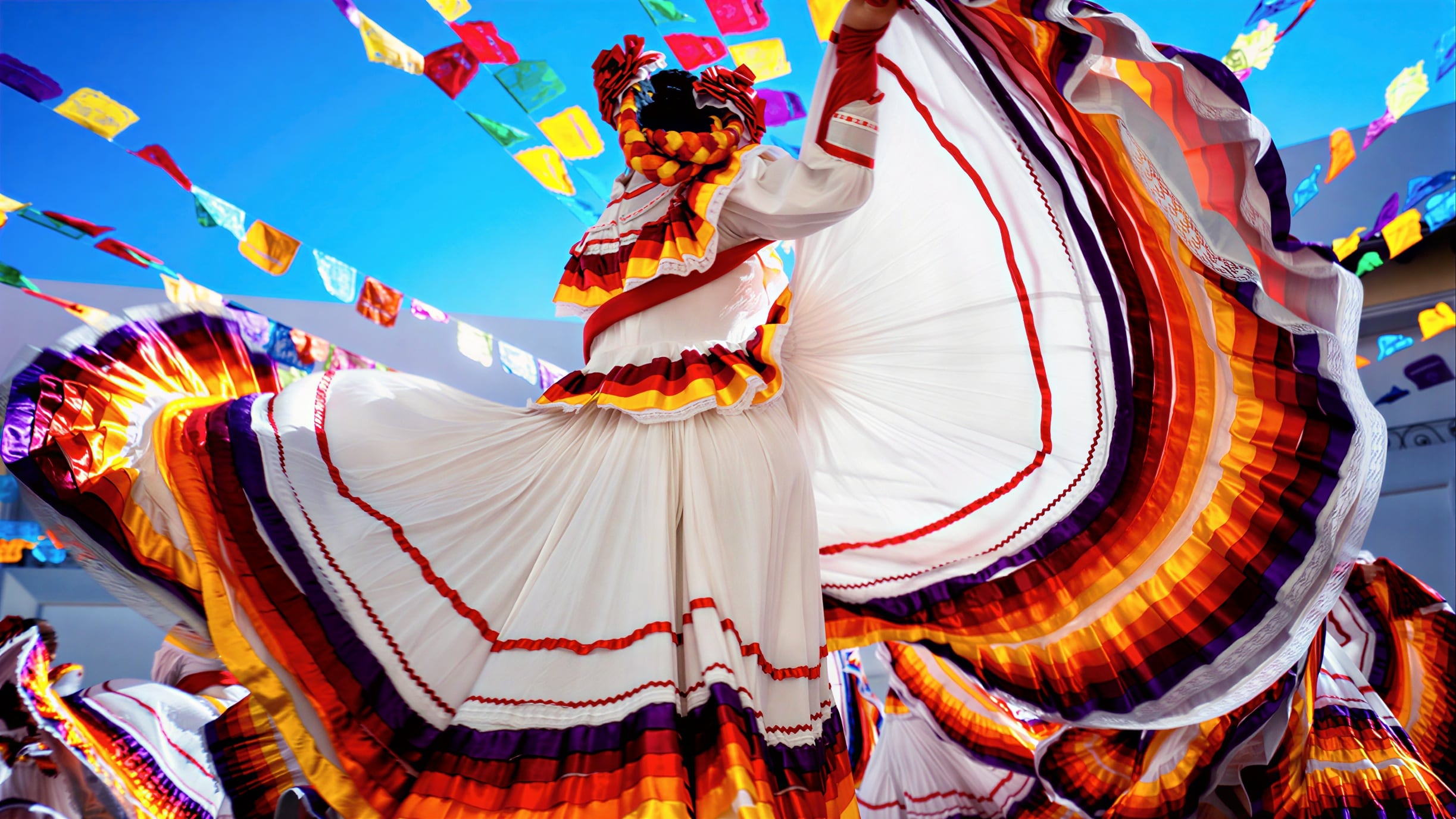 A dancer in a bright traditional dress twirls fabric under colorful flags against a blue sky.