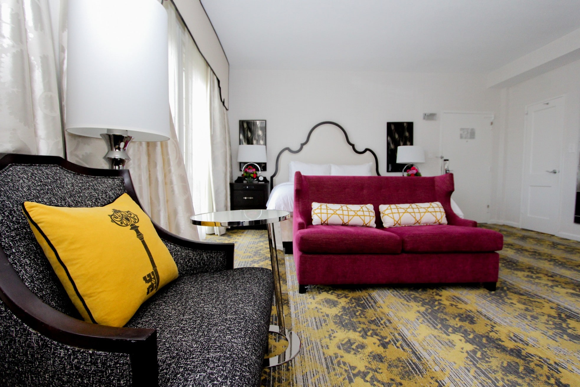 A hotel room with a purple sofa, a patterned chair, and a bed in the background.