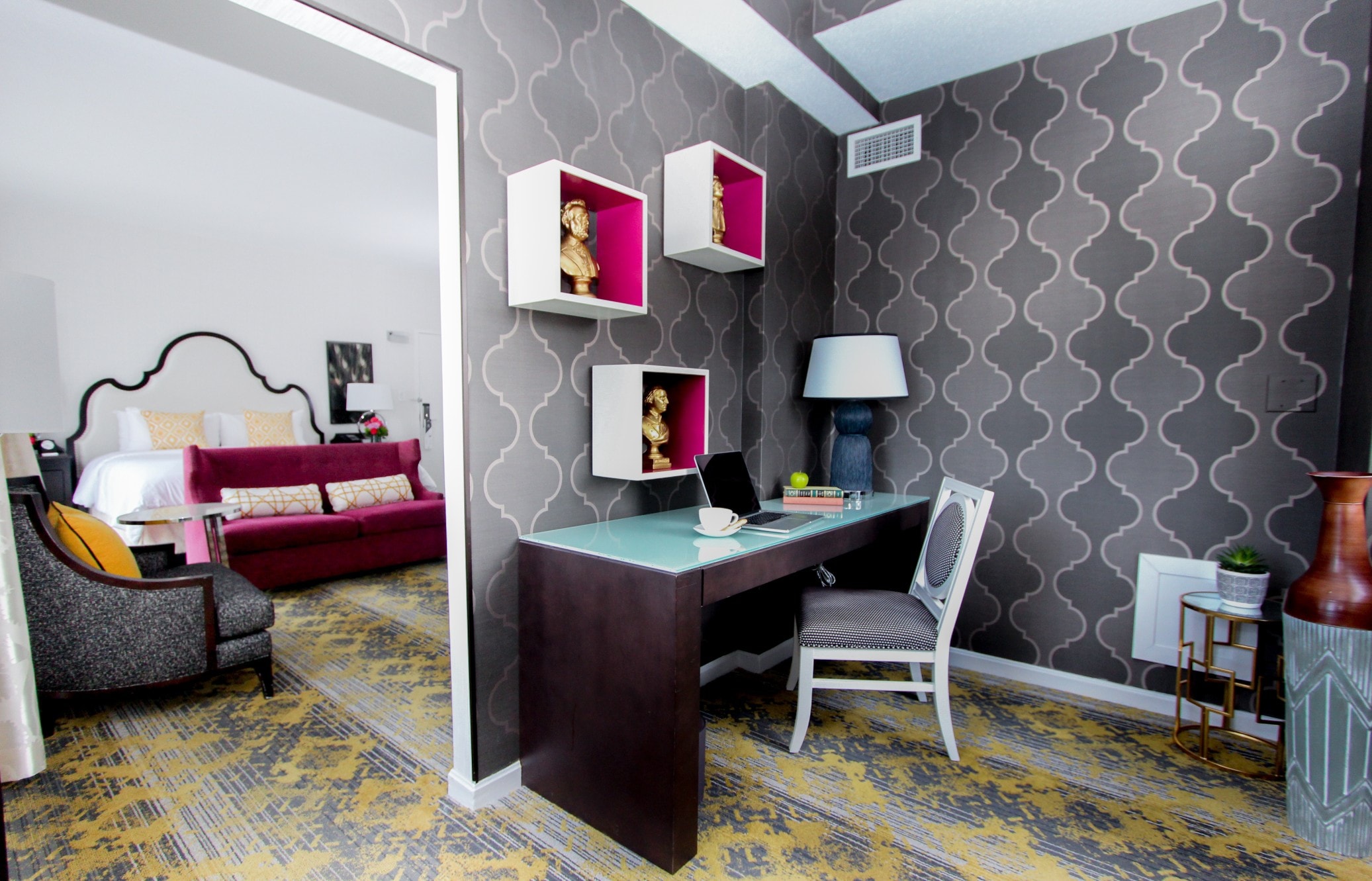 A hotel room with a desk, chair, and shelves, with a bed and couch in the background.