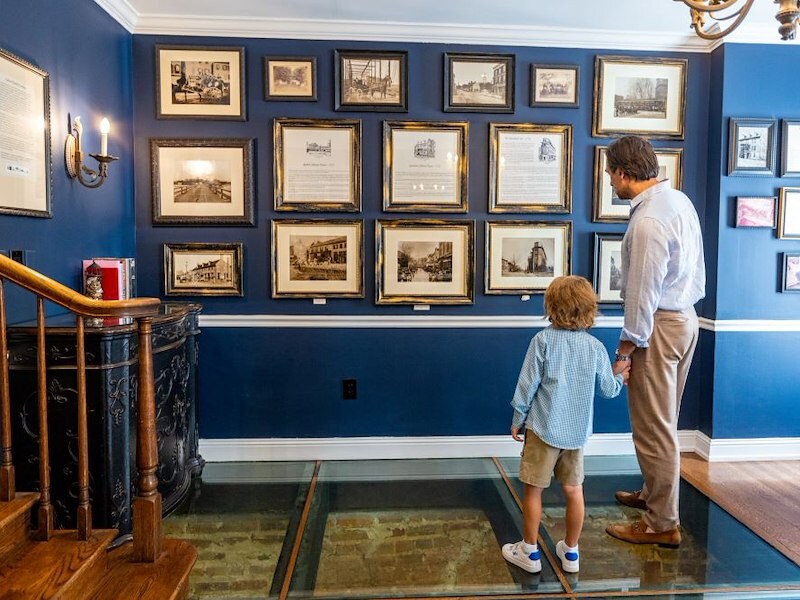 Father and son viewing framed historical photos and documents on a blue wall inside a heritage room.