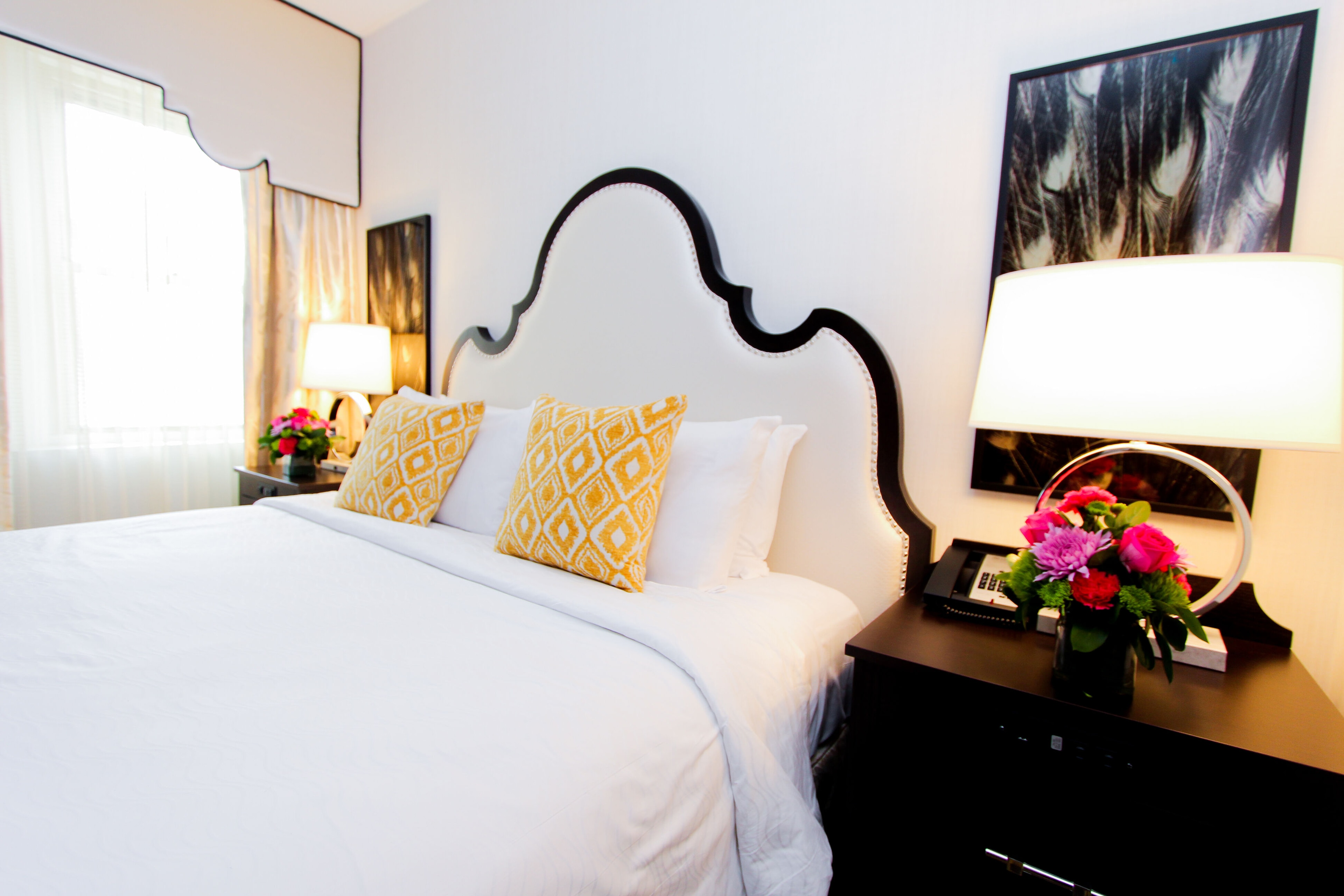 A neatly made bed with white linens and yellow pillows in a room with a large headboard and a window with sheer curtains.