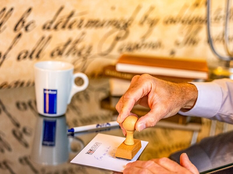 Hand stamping a postcard on a desk with books, pen, and coffee mug at Melrose Georgetown Hotel.