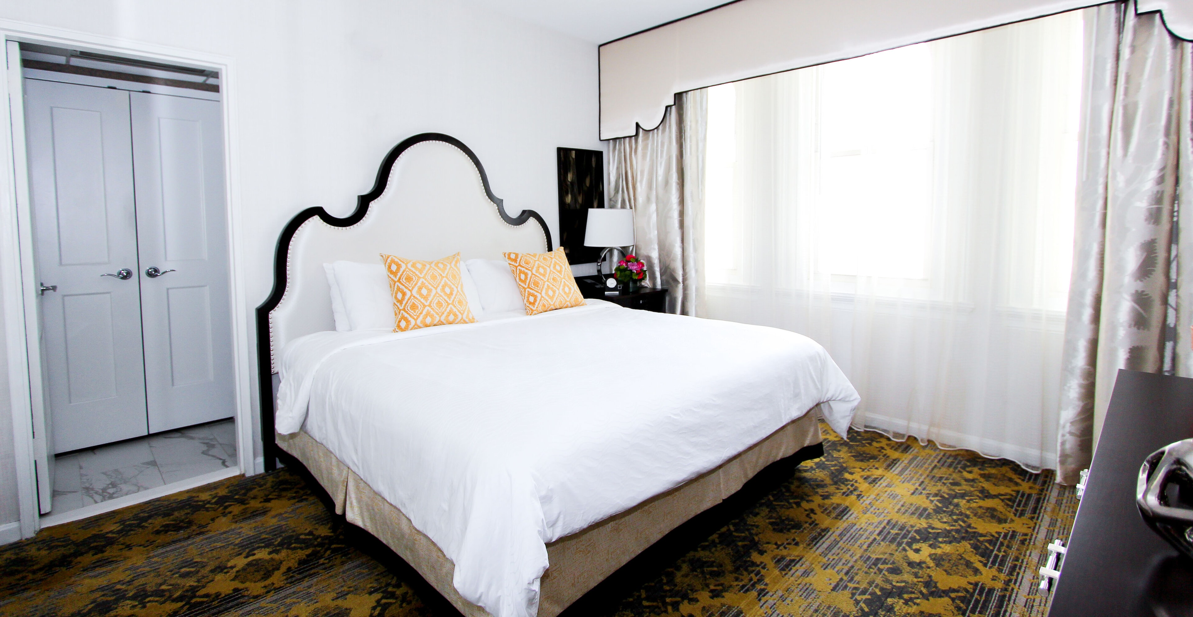 A wide-angle view of a hotel room with a large bed, yellow and black carpet, and a window with sheer curtains.