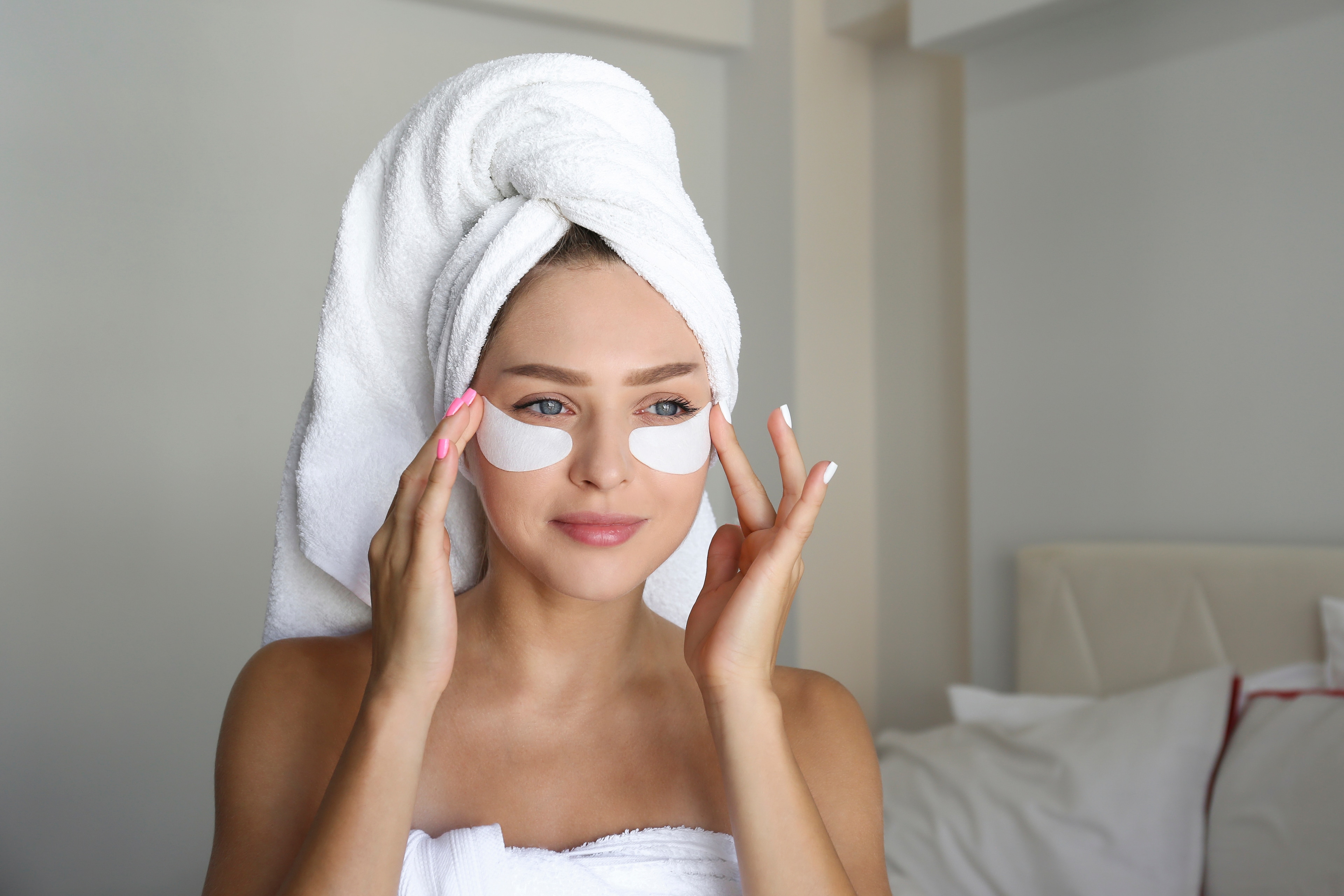 A woman in a towel with eye patches on her face in a bedroom