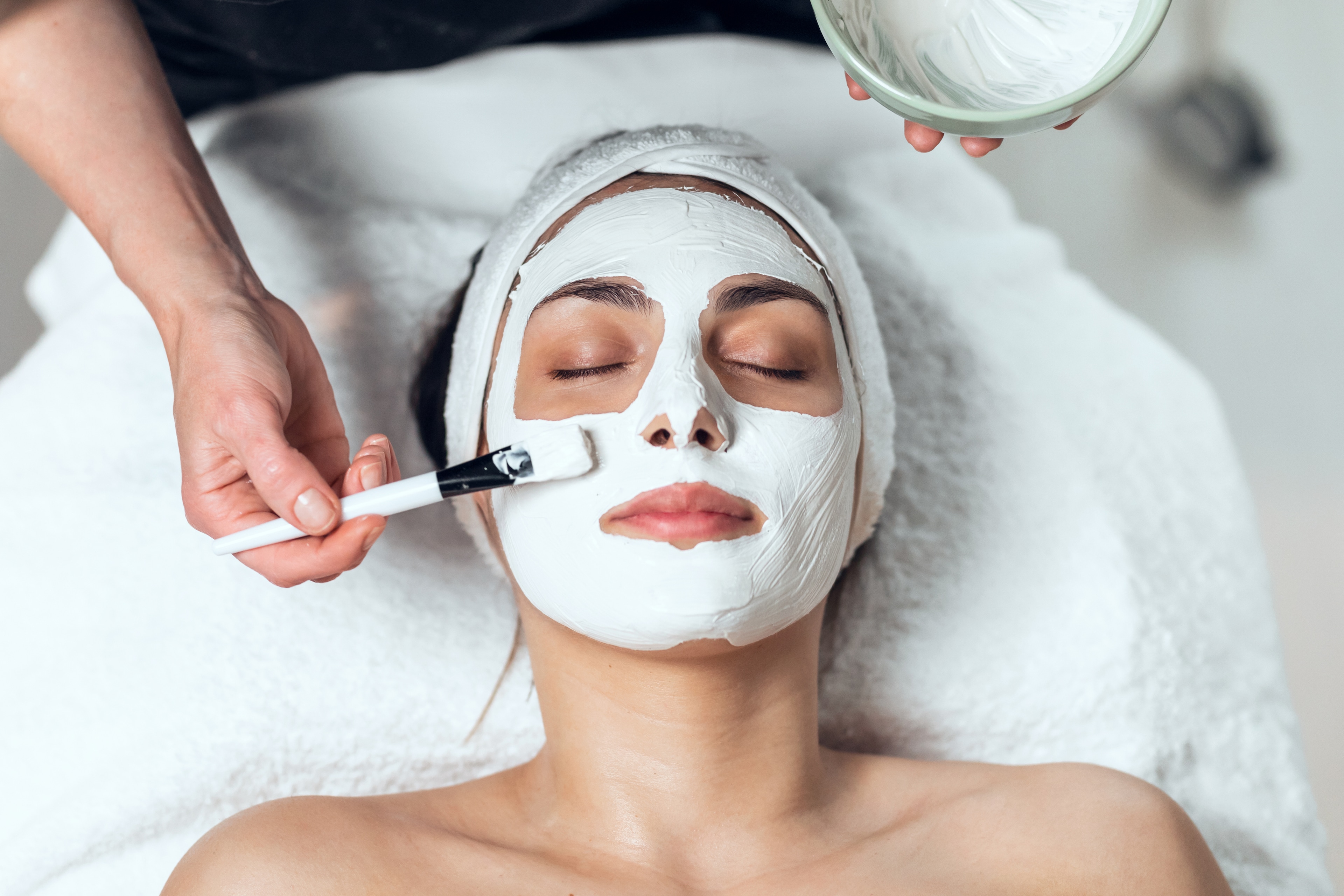 A woman receiving a facial treatment with a white mask being applied to her face.