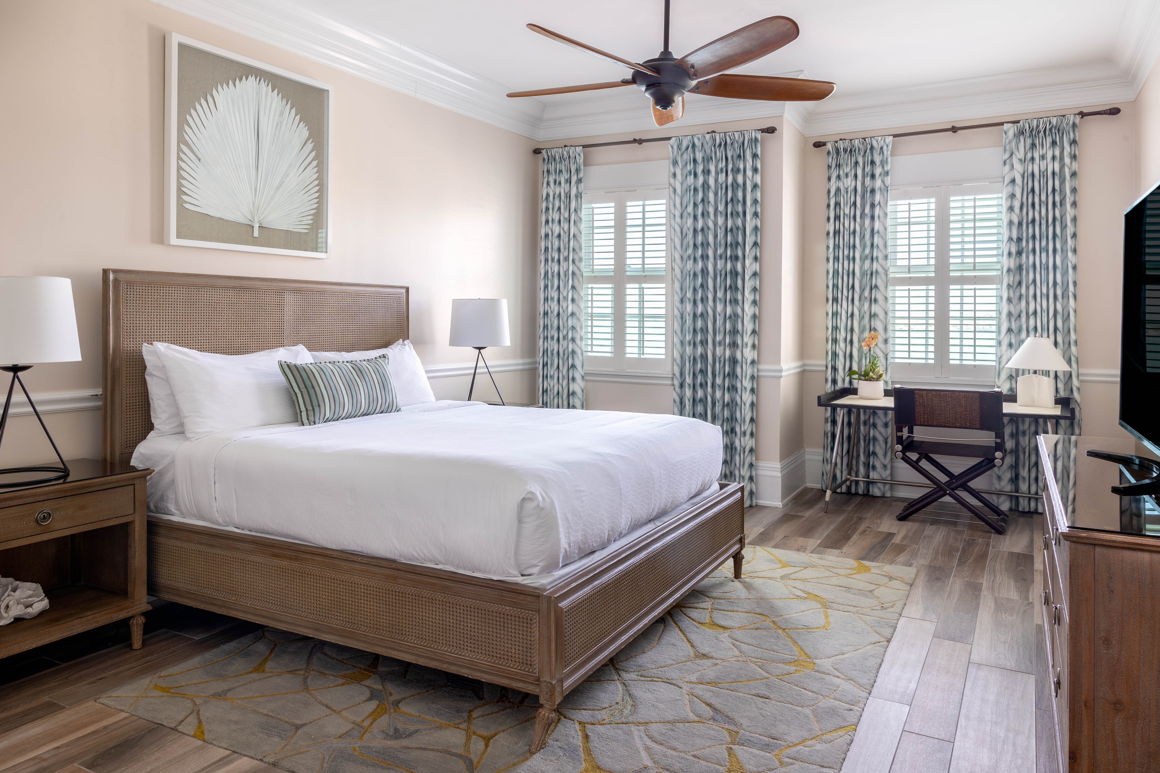 A wide view of a guest room with a bed, nightstand, lamp, ceiling fan, window, and desk.