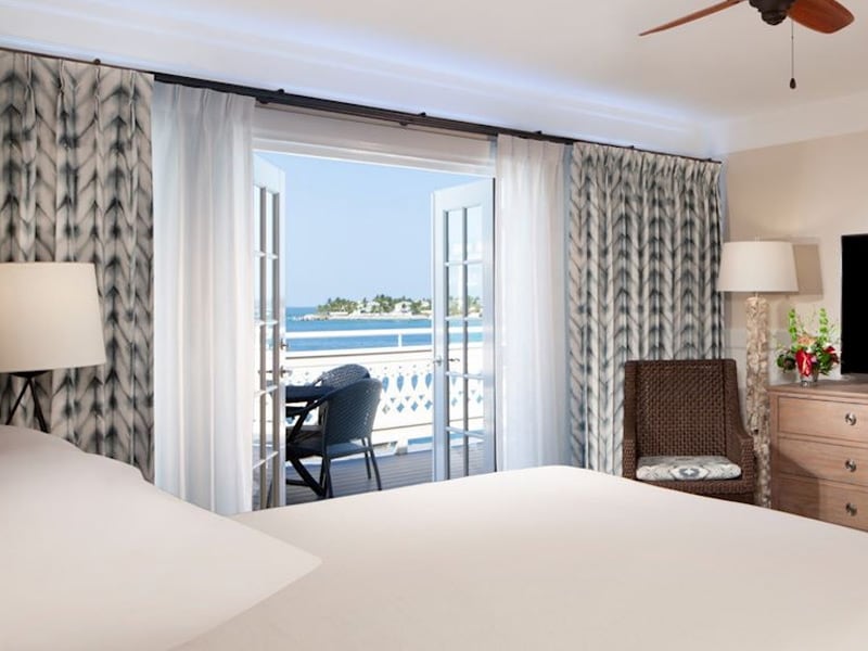 A wide-angle view of a hotel room with a large bed, dresser, wicker chair, and glass doors leading to a balcony with a view.