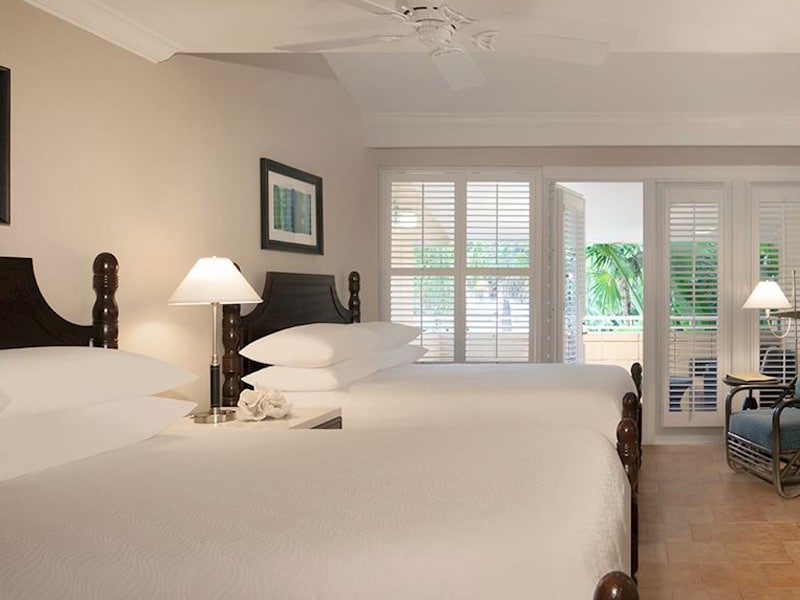 A well-lit hotel room with two beds, a ceiling fan, and a large window with blinds.