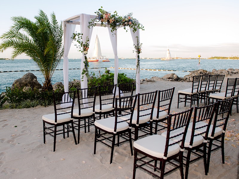 A beachside wedding setup with a floral arch, white chairs, and a palm tree, overlooking sailboats on the water.