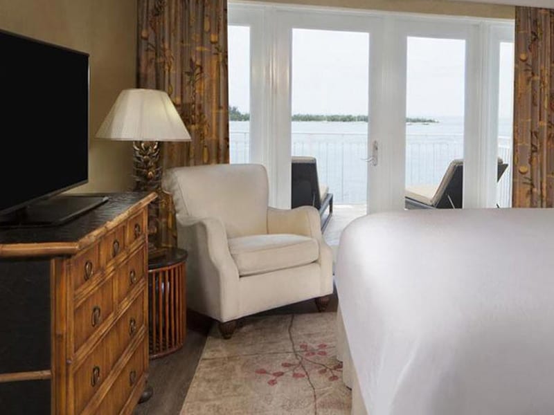 A hotel room with a white bed, armchair, TV, and lamp, with a view of the water through a glass door.