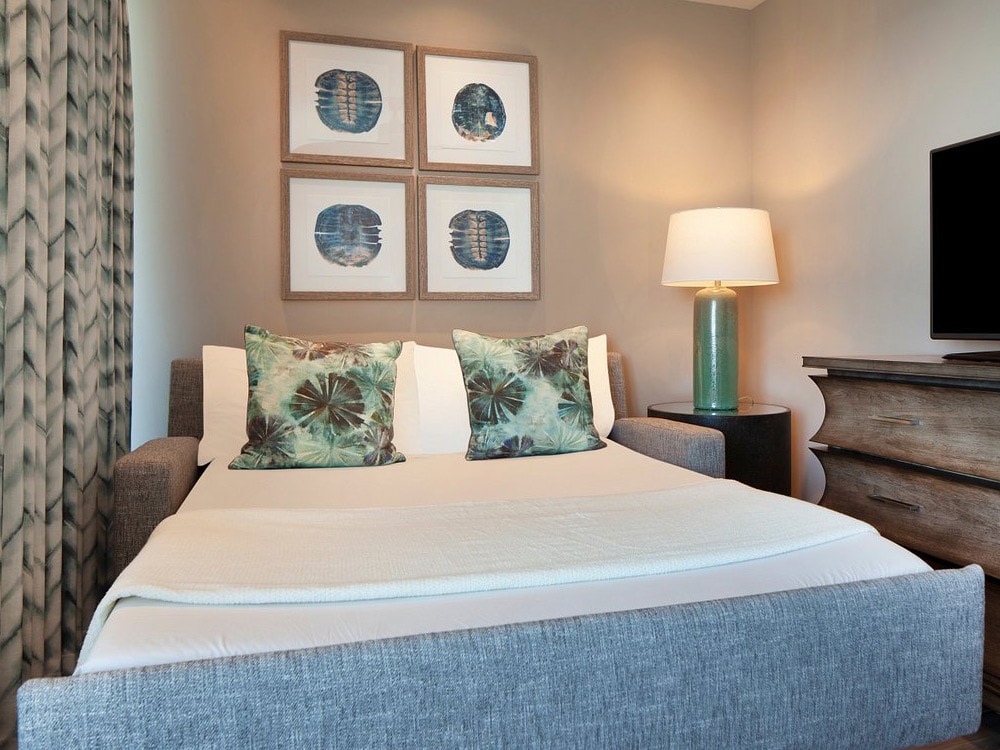 A neatly made bed with white linens and green pillows in a room with a lamp, TV, and artwork on the wall.