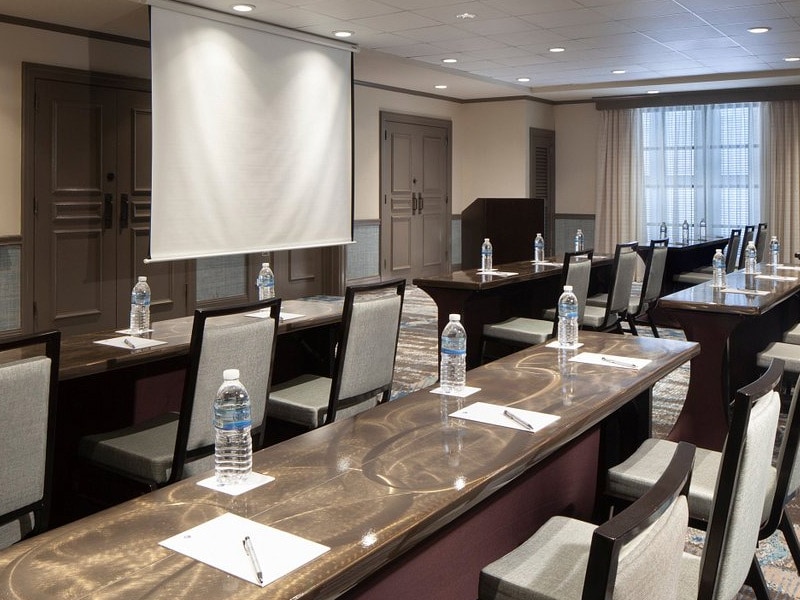 A meeting room with a long dark wood table, gray chairs, a projector screen, and a large window.