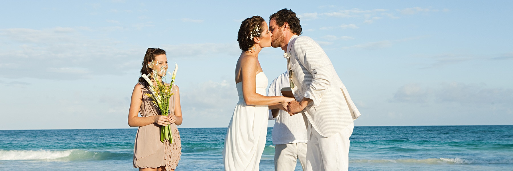A couple in wedding attire kisses on the beach with a girl holding flowers nearby.