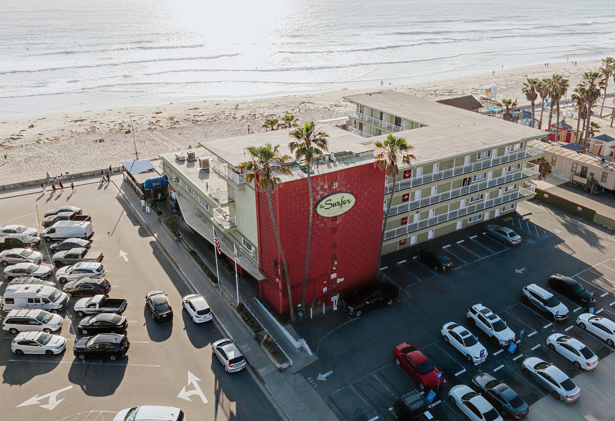 An aerial view of a beachfront hotel with a red and white building, a parking lot, and palm trees.