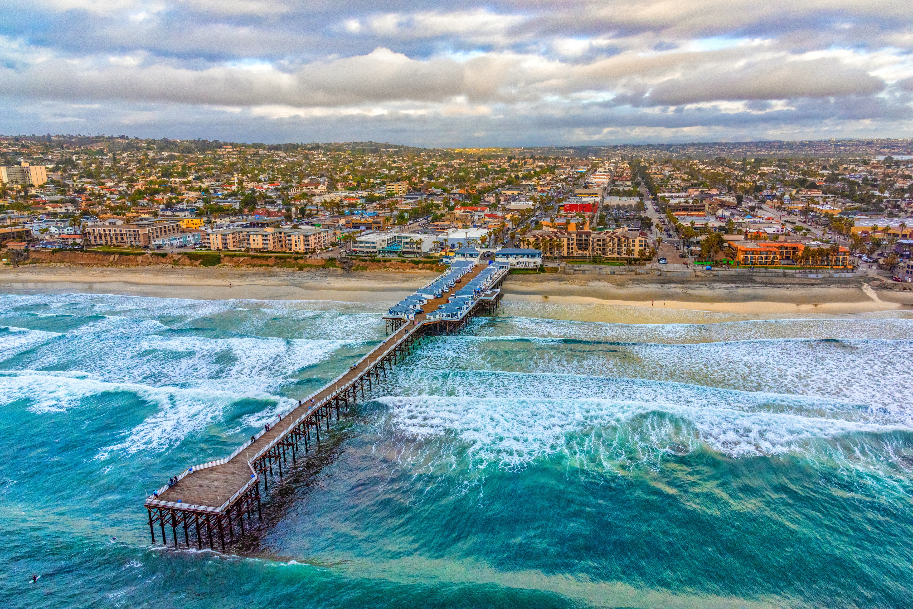 Aerial view of a long pier extending into the ocean with waves crashing against it, in front of a beach and buildings under a cloudy sky.