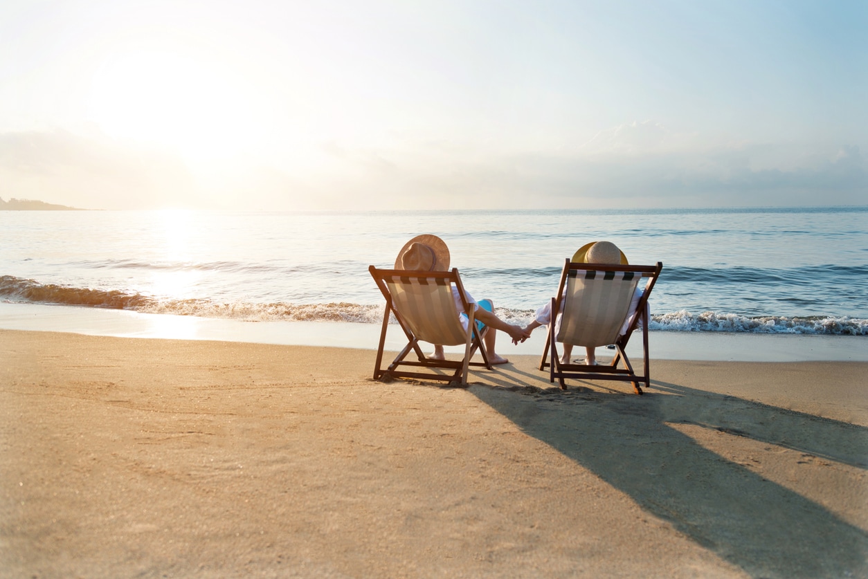A couple sitting on beach chairs, holding hands, and looking out at the ocean during sunset.