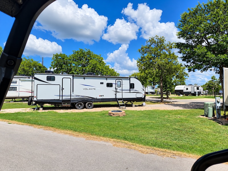 White RV parked at Great Escapes Austin Oaks, surrounded by green lawns and shady trees under a blue sky.