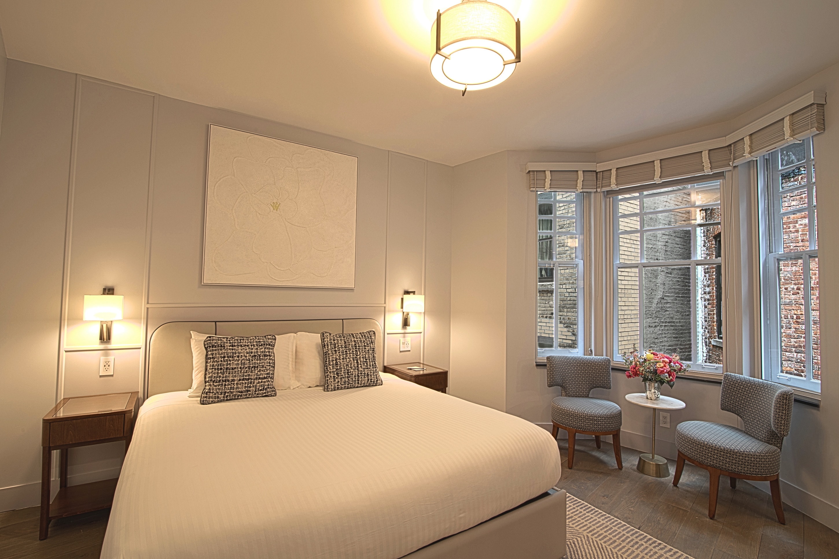A neatly made hotel room with a king-size bed, two nightstands with lamps, a window with a view, and two chairs with a small table.