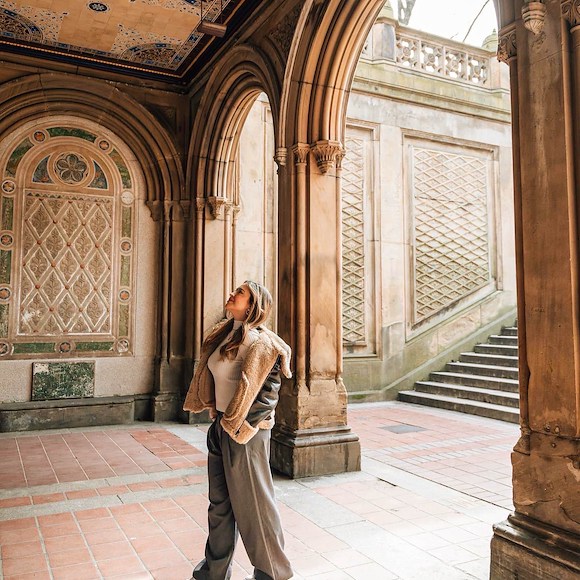 A woman stands in a historic, ornate outdoor space with arches, columns, and a tiled floor.