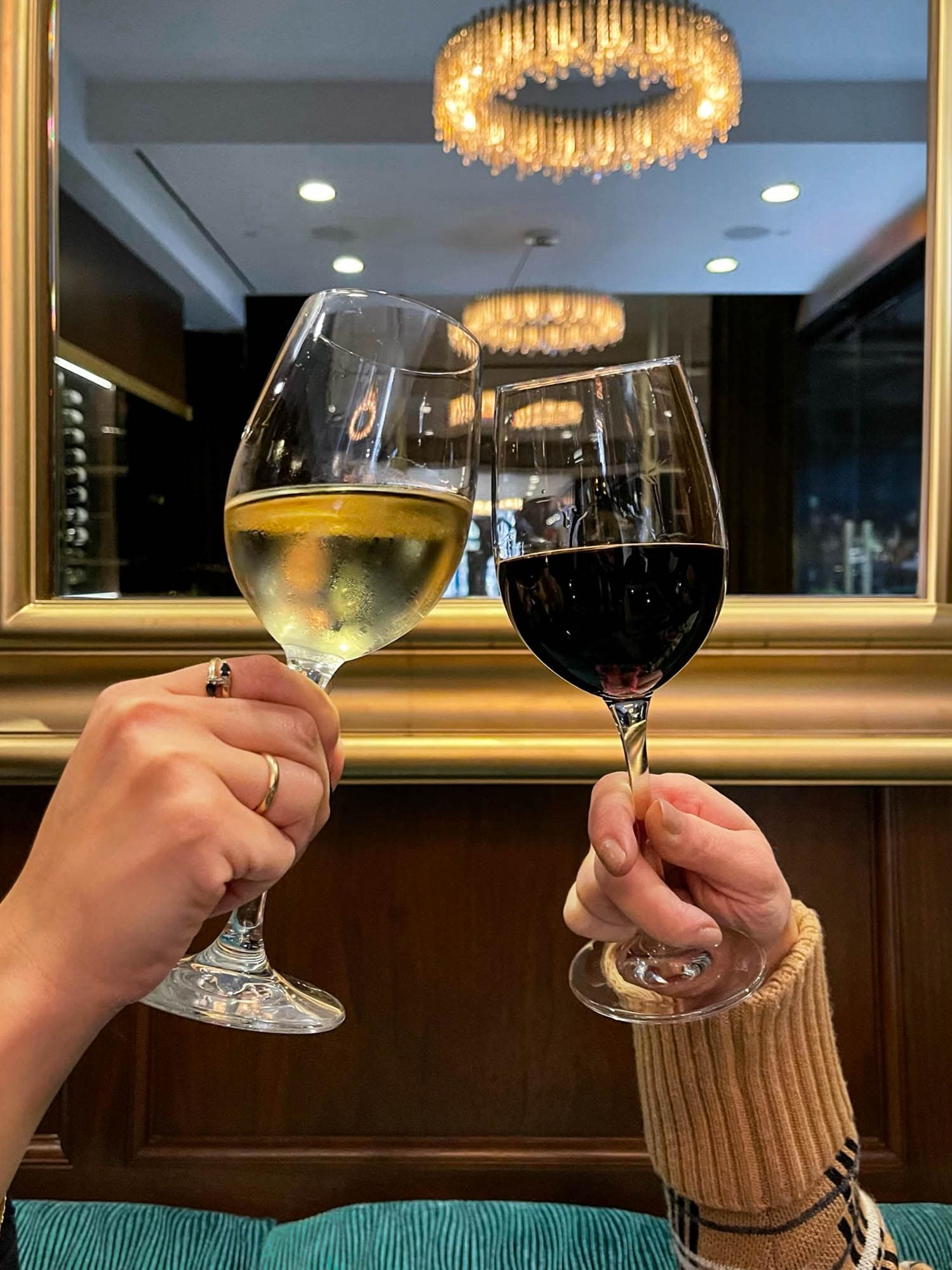 Two hands holding wine glasses in front of a mirror with a gold frame and a chandelier in the background.