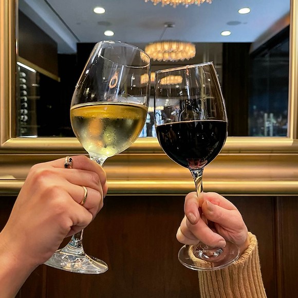 Two hands holding wine glasses in front of a mirror with a gold frame and a chandelier in the background.
